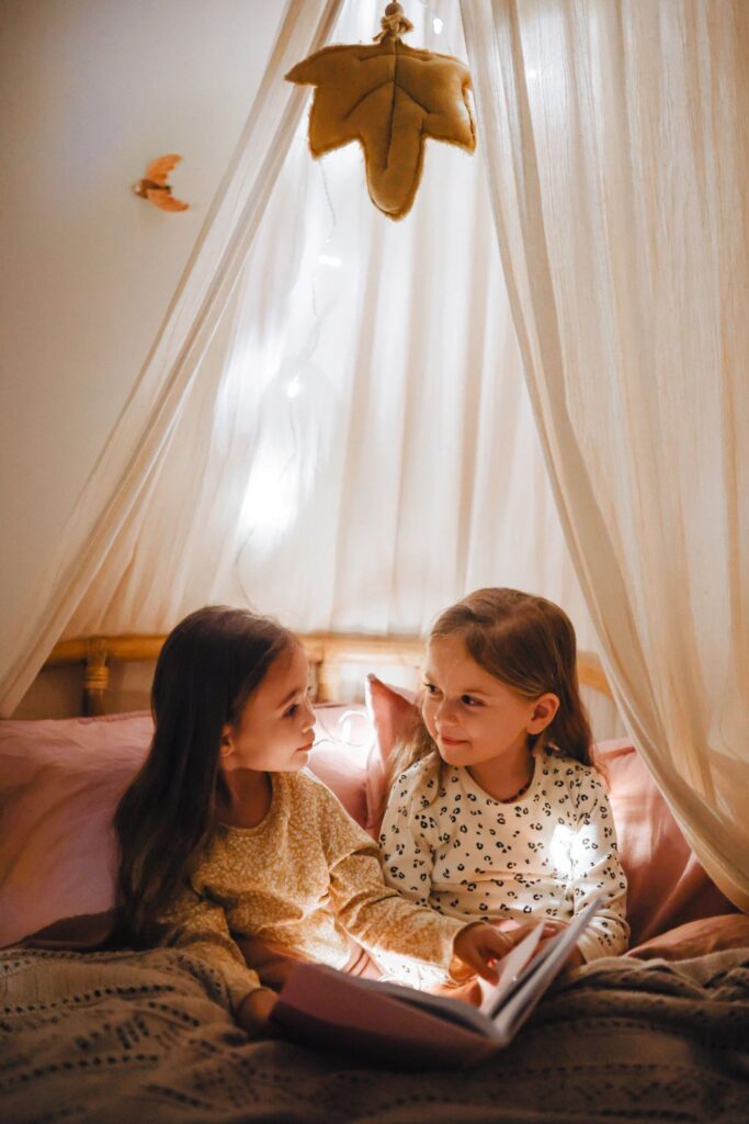 Two cute sisters are sitting on the bed and reading a funny book, smiling and laughing,