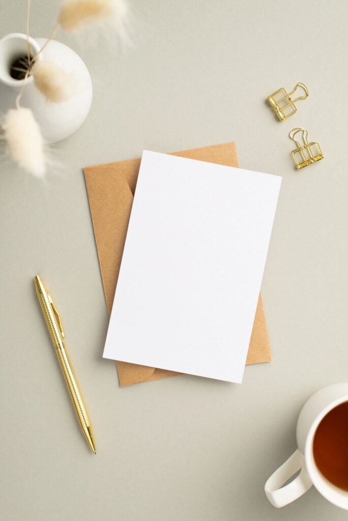 Business concept. Top view vertical photo of paper sheet craft paper envelope gold pen binder clips cup of tea and vase with white lagurus flowers on pastel grey background with copyspace
